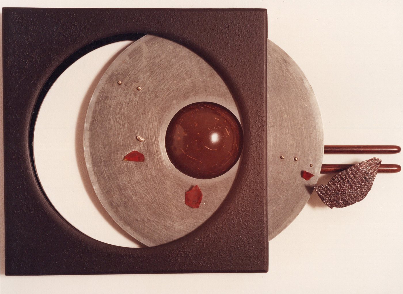 Broche van lange speelduur, 1978 - a large pale wooden disc on a dark brown square, a deep red sphere set into its centre, with small red fragments and a woven form at the right.