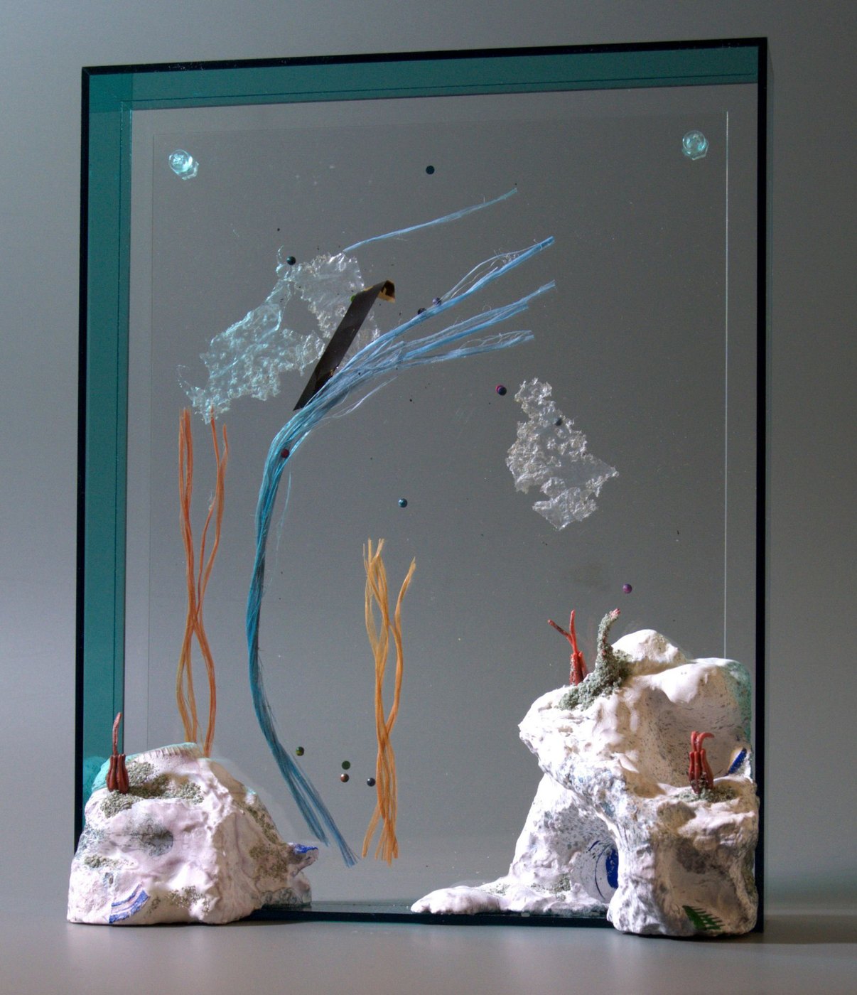 Plastiec, 2026 - a standing glass panel in a green-edged frame, with blue and orange strands, crinkled clear plastic and small coloured beads on the glass, between two white rock forms with red coral-like sprigs.