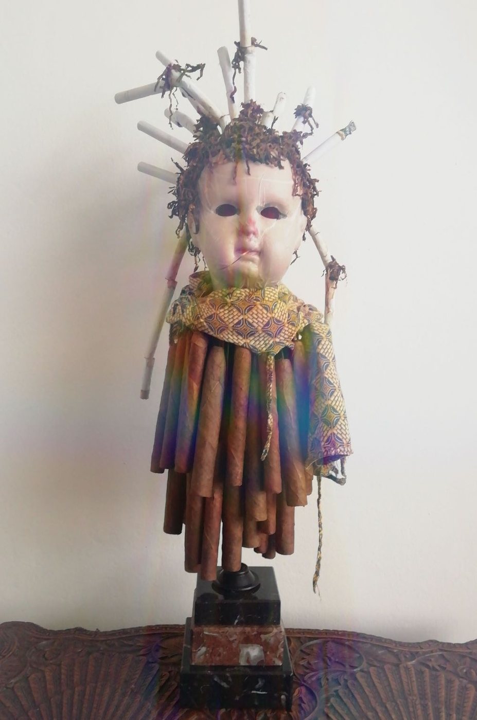 Totem Smokem, 2024 - a doll figure in a ruff and striped robe, its head crowned with a burst of cigarettes and thin sticks.